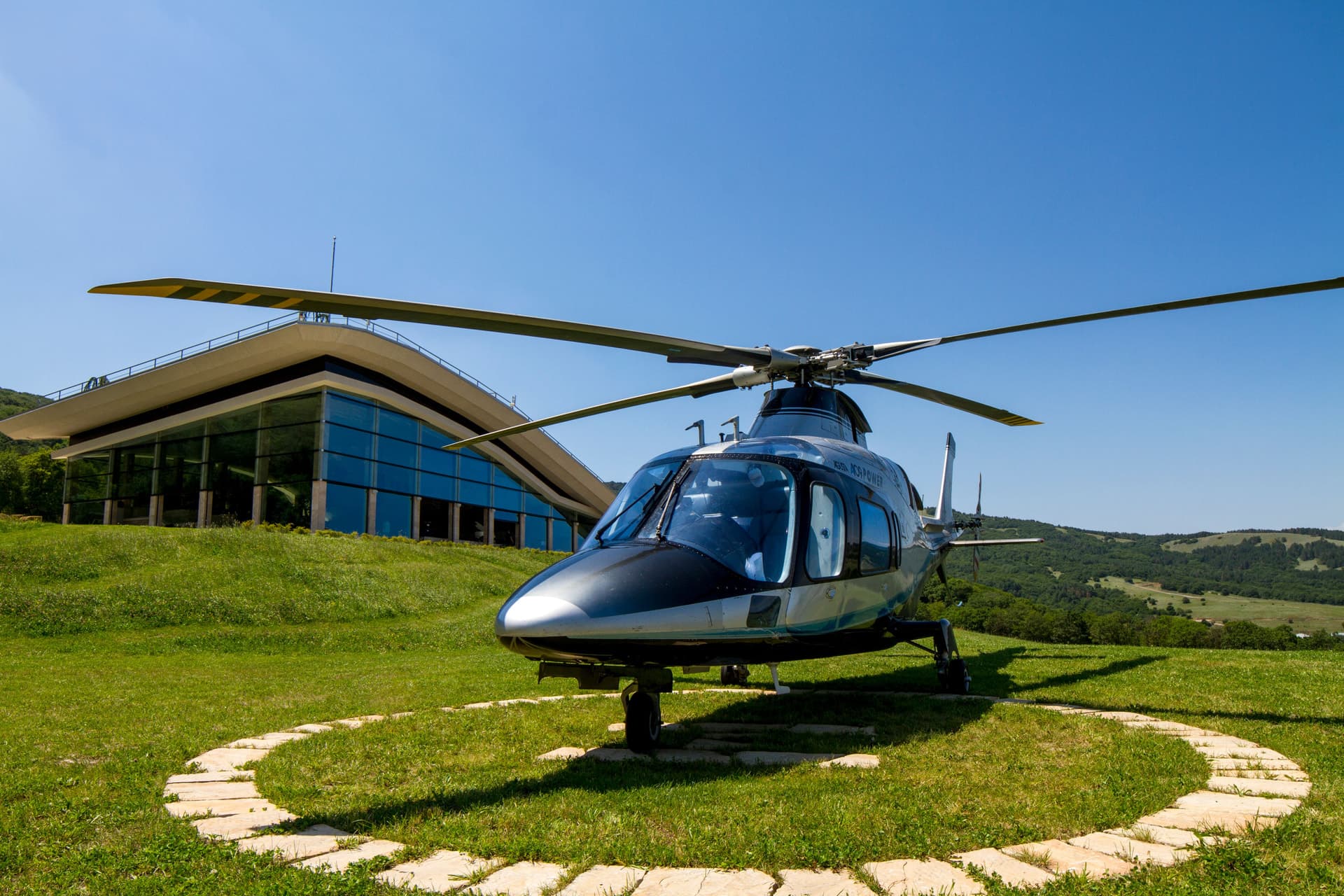 Helicopter on grass beside a glass-fronted building and green hills